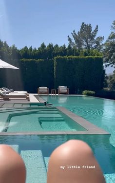 someone's feet in the water next to a swimming pool with lounge chairs and an umbrella