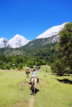 two people riding horses on a grassy field with mountains in the backgrouds