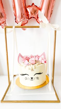 a white cake with pink flowers and cat ears on top, surrounded by foil balloons