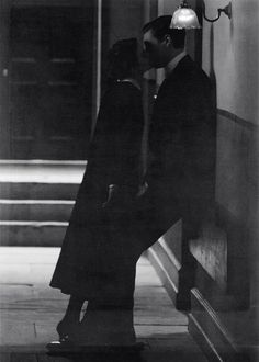 black and white photograph of man kissing woman on the cheek in front of building with stairs