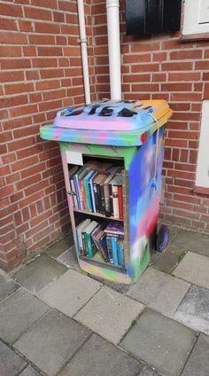 a multicolored trash can sitting next to a brick wall with books on it