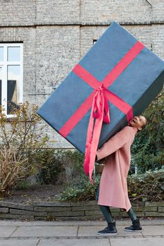 a woman carrying a large blue and red gift box on her back while standing in front of a brick building