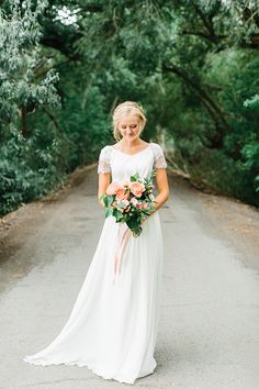 a woman in a white dress is standing on a road with trees and bushes behind her