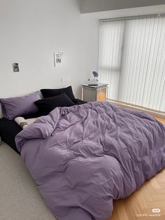 an unmade bed with purple sheets and black pillows on the floor in front of a window