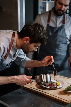 two men in aprons are preparing food on a cutting board and one man is holding a spoon