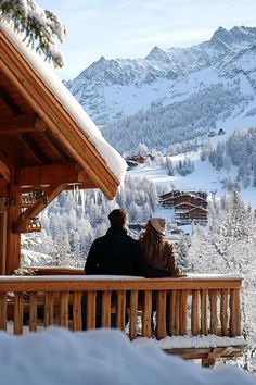 two people sitting on a bench in front of a snowy mountain side area with trees and buildings