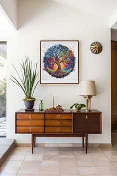 a painting hangs on the wall next to a dresser with two potted plants and a lamp