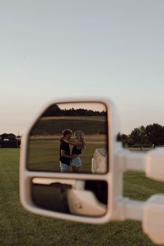 two people standing in the grass looking at their reflection in a rear view mirror with trees in the background