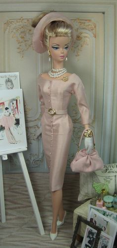 a barbie doll in a pink dress and hat next to a white easel with pictures on it