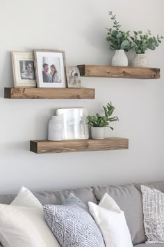 three wooden shelves on the wall above a gray couch with pillows and plants in vases