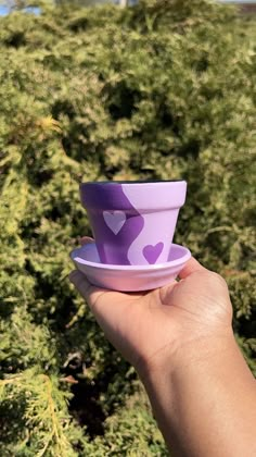 a hand holding a purple cup with hearts on it in front of some green bushes