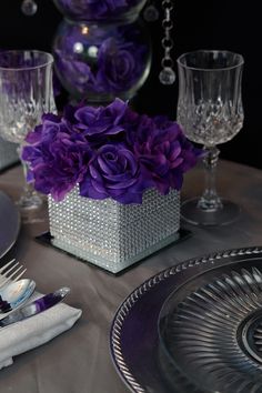 purple flowers are in a square vase on a table set with silver plates and crystalware