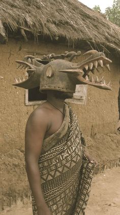 a man with a mask on his head standing in front of a mud hut