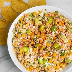 a white bowl filled with corn salad next to tortilla chips on a marble surface