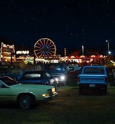 there are many cars that are parked in the parking lot at night time, with ferris wheel in the background