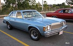 an old blue car parked in a parking lot next to another red car on the street