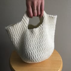 a hand holding a white knitted bag on top of a wooden table next to a wall