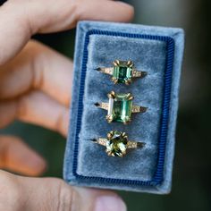 three rings with green and yellow stones are in a blue velvet ring box that is being held by someone's hand