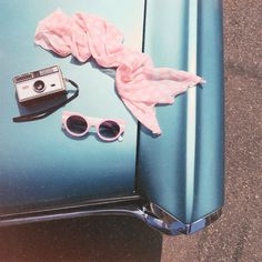 an old blue car with sunglasses and a pink scarf on it's hood next to a polaroid camera