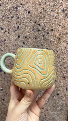 a hand holding a yellow and blue coffee cup in front of a carpeted wall