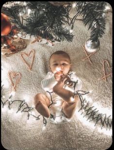 a baby sitting under a christmas tree surrounded by ornaments and lights on the floor, looking up at the camera