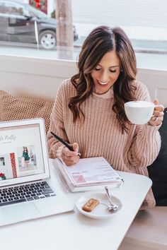 a woman sitting at a table in front of a laptop holding a cup and writing