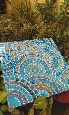 a blue and gold patterned piece of art sitting in front of some potted plants