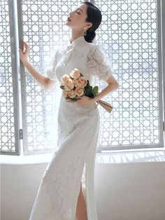 a woman in a white dress is standing near a window and holding a flower bouquet