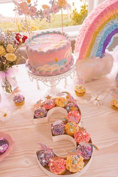 a table topped with a cake and cupcakes next to a rainbow shaped cake