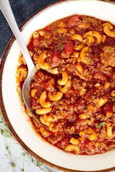 a white bowl filled with pasta and meat on top of a marble counter next to a silver spoon