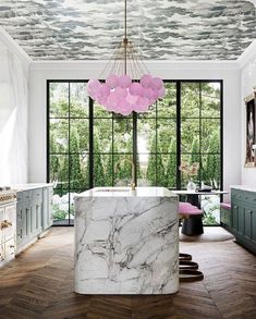 a marble island in the middle of a kitchen with green cabinets and large windows that look out onto trees