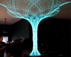 an illuminated tree in the middle of a room with people standing around and looking at it