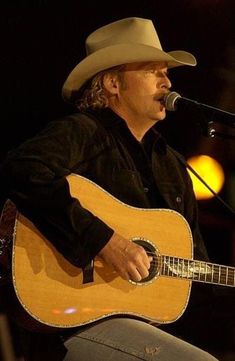 a man wearing a cowboy hat and holding a guitar in front of a microphone on stage