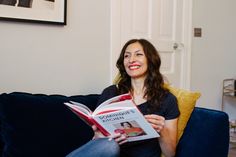 a woman sitting on a blue couch reading a book with her legs crossed and smiling at the camera