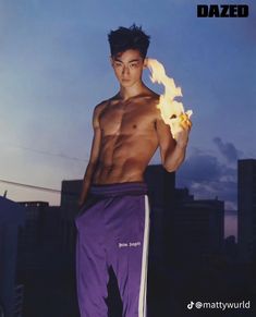 a shirtless man holding a fireball in front of his face with buildings in the background