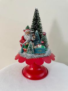 a small christmas tree on top of a red stand with santa clause and other decorations