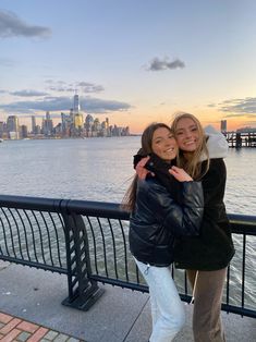 two women standing next to each other in front of a body of water with a city skyline behind them