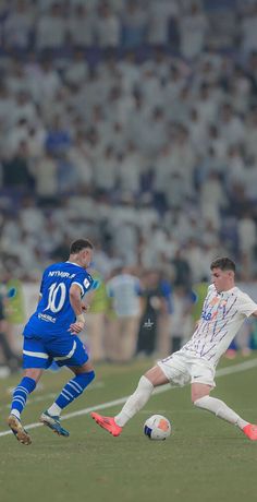 two men playing soccer on a field with people watching from the stands in the bleachers