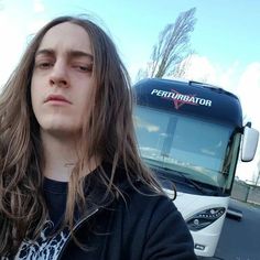 a man with long hair standing in front of a bus and looking at the camera