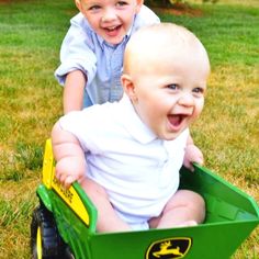 two young boys are sitting in a green toy truck on the grass and one boy is smiling