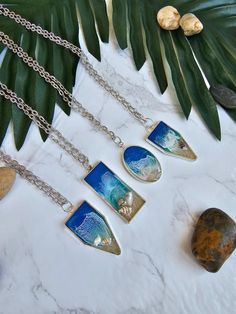 five different necklaces on a table with leaves and rocks in the background, including one featuring an ocean scene