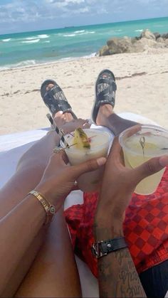 two people sitting on the beach with drinks in their hands and one person wearing flip flops