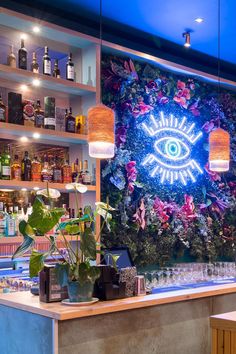 a bar with plants and bottles on the wall next to an eye - shaped neon sign