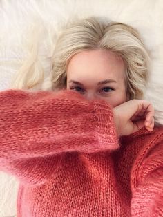 a woman laying on top of a bed wearing a pink sweater and holding her hand under her chin