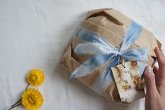 a hand is holding a wrapped present with blue ribbon on it and yellow flowers next to it