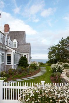 a white picket fence in front of a house with flowers on the lawn and water behind it