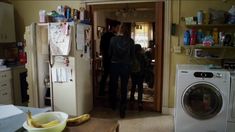 two people standing in a kitchen next to a washer and dryer with the door open