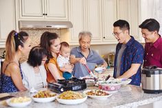 a group of people standing around a kitchen counter with food on the table and an adult holding a baby