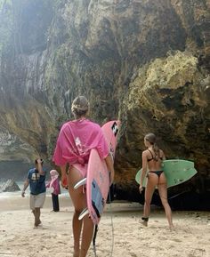 two women in bikinis carrying surfboards on the beach with large rocks behind them