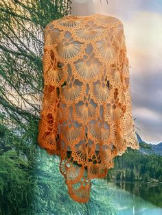 an orange crocheted shawl hanging from a tree next to a body of water